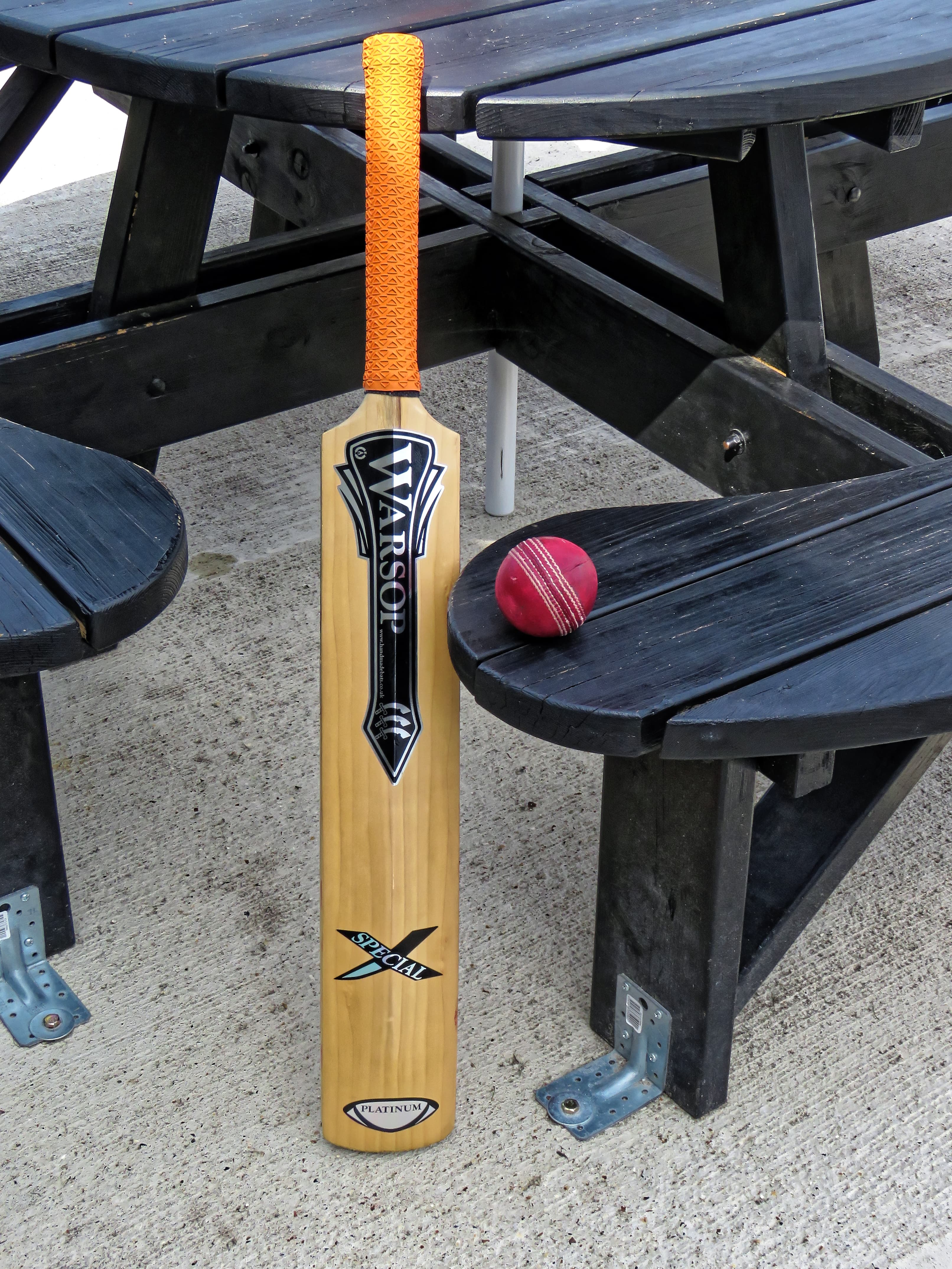 A cricket bat with an orange grip and a red cricket ball beside a wooden bench